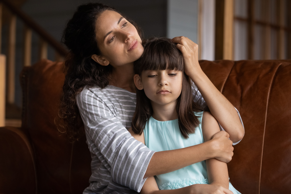 Close up caring mother hugging, calming upset little daughter, sitting on couch at home, mum comforting offended sad preschool girl, expressing love and support, child psychologist concept
