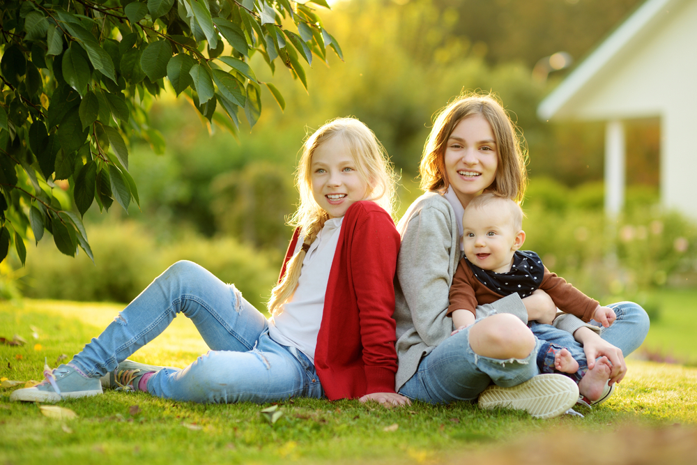 Two big sisters and their infant brother having fun outdoors. Two young girls holding their baby boy sibling on summer day. Kids with large age gap. Big age difference between siblings. Big family.