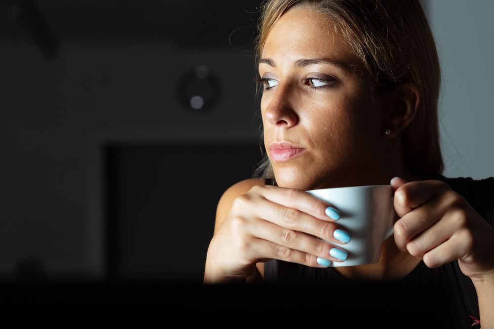 Thoughtful woman sitting working on a laptop in the darkness holding a mug of coffee in her hand as she looks of to the side in close up