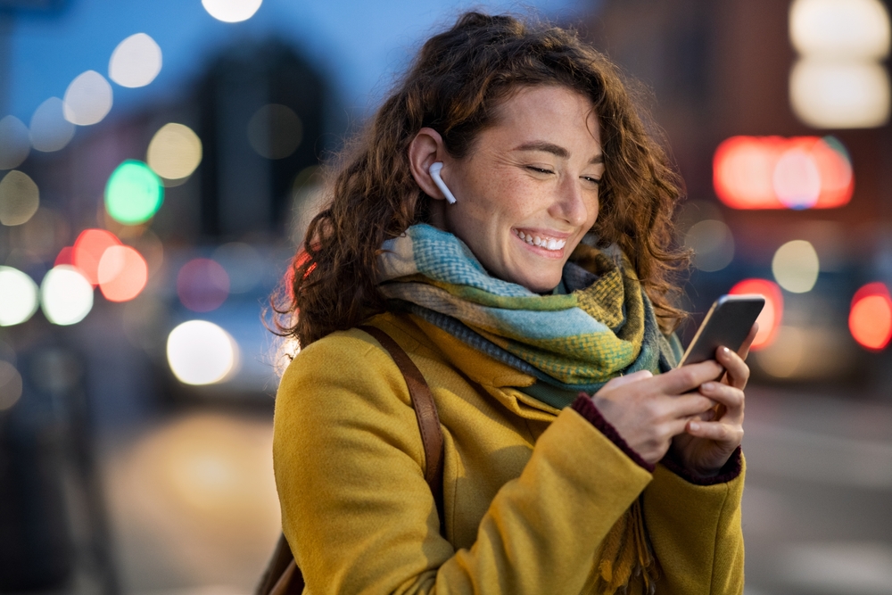 Beautiful woman using mobile phone while listening voice note with wireless earbuds. Smiling girl changing songs from smart phone in a city street. Happy smiling woman chatting on smartphone in winter