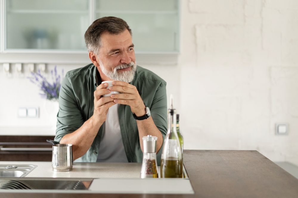 Happy mature man drinking coffee at home in the kitchen, enjoying hot drink in the morning on weekend