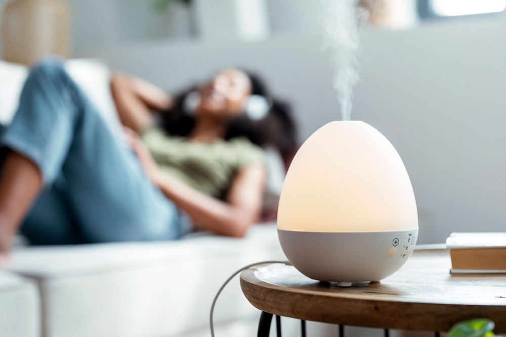 Shot of essential oil aroma diffuser humidifier diffusing water articles in the air while woman listening music lying on coach.