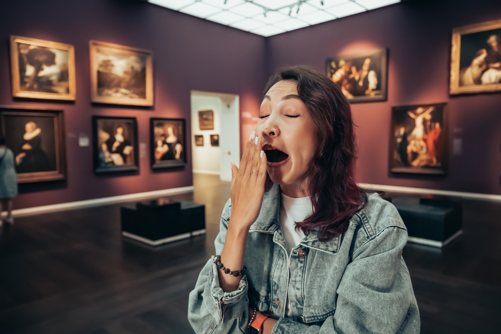 a bored girl visitor or student in an art gallery or museum yawns and looks at the masterpieces of classical artists
