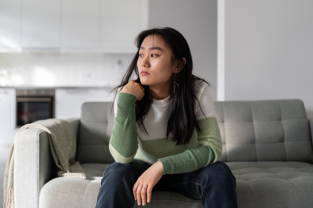 Sad lonely young Asian woman having depression symptoms, sitting on sofa at home looking into distance, upset unhappy female worrying about money or family problems. Mental disorders,