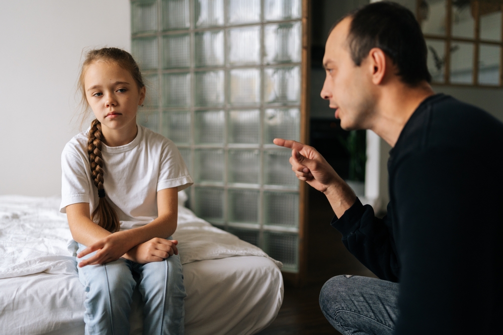 Portrait of loving young father having serious conversation with naughty adorable little daughter , talking about rules to her sitting on bed at home. Concept of dad-daughter problem in preteen years.