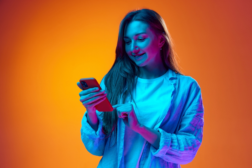 Smiling young woman typing text message on mobile phone over gradient orange studio background in neon light. Online communication. Concept of youth, human emotions, lifestyle, facial expression, ad
