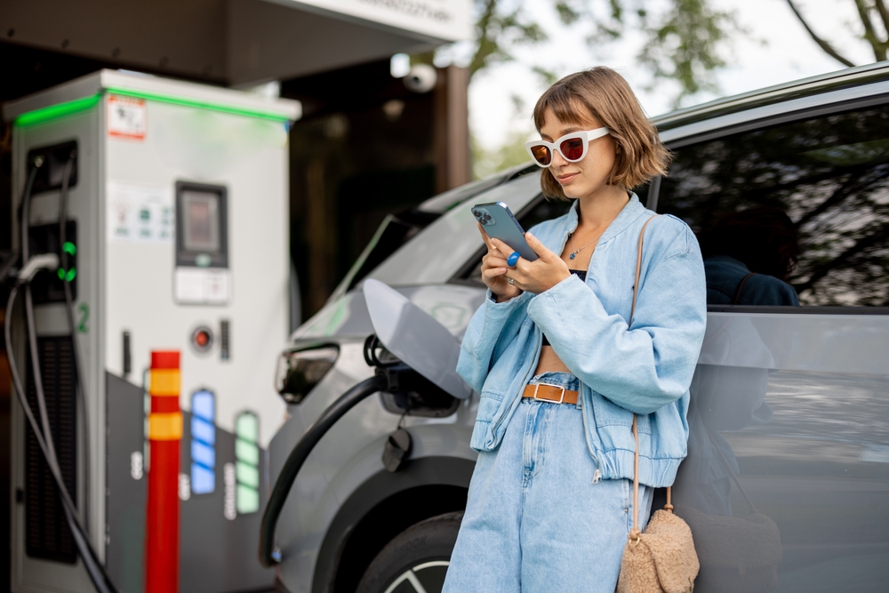 Young woman using a smart phone, while waiting for her electric car to charge on a public charging station outdoors. Concept of modern technologies and lifestyle