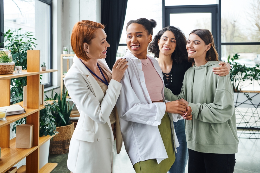 pleased psychologist looking at overjoyed african american woman near multiethnic girlfriends during motivation session in consulting room, female unity and support concept
