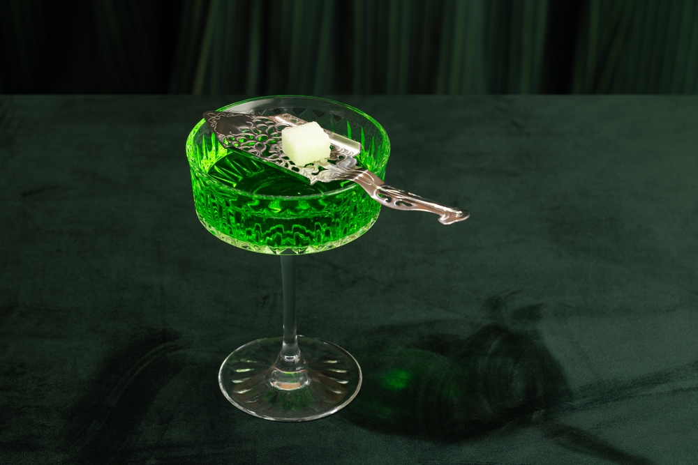 Green cocktail in a crystal glass with a sugar cube on a silver spoon on a dark green background
