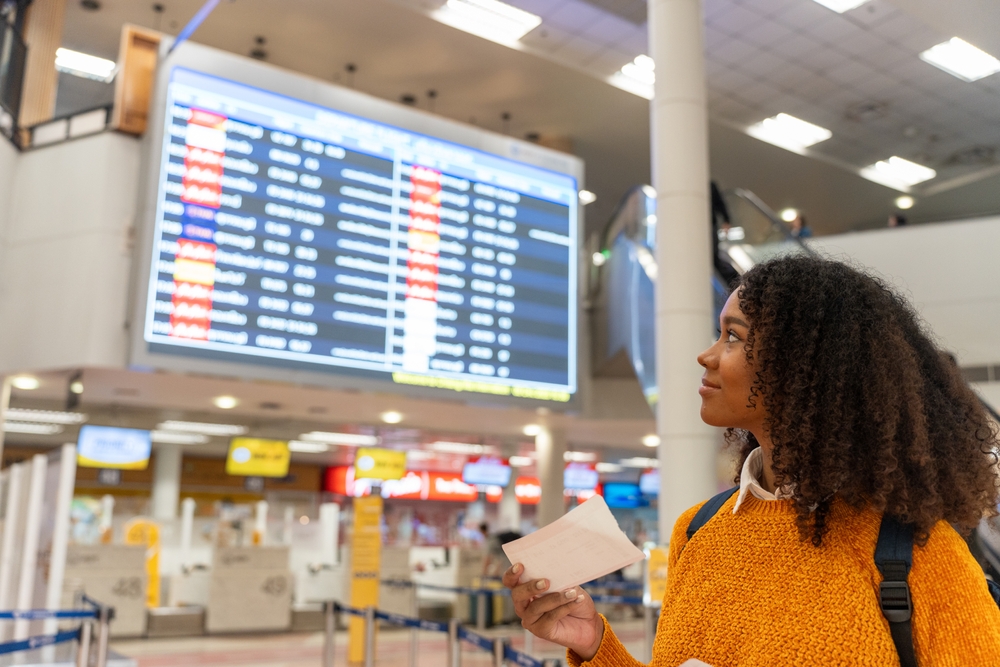 Black woman looks at the flight schedule on a digital monitor in an airport to check the gate and time to board the plane to travel or study abroad.