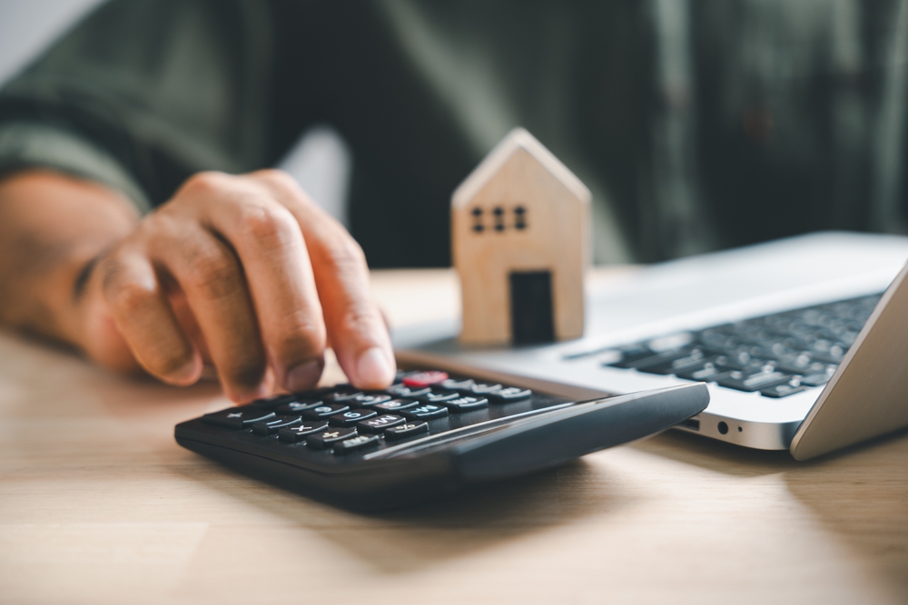 Pressing calculators hand eyes home refinance. Wooden house model, buy or rent note on desk. Saving for property purchase concept efficient mortgage payment. Tax credit analysis for financial strategy
