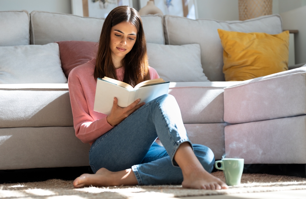 Shot of pretty young woman reading a book and drinking a cup of coffee while sitting on the floor at home.