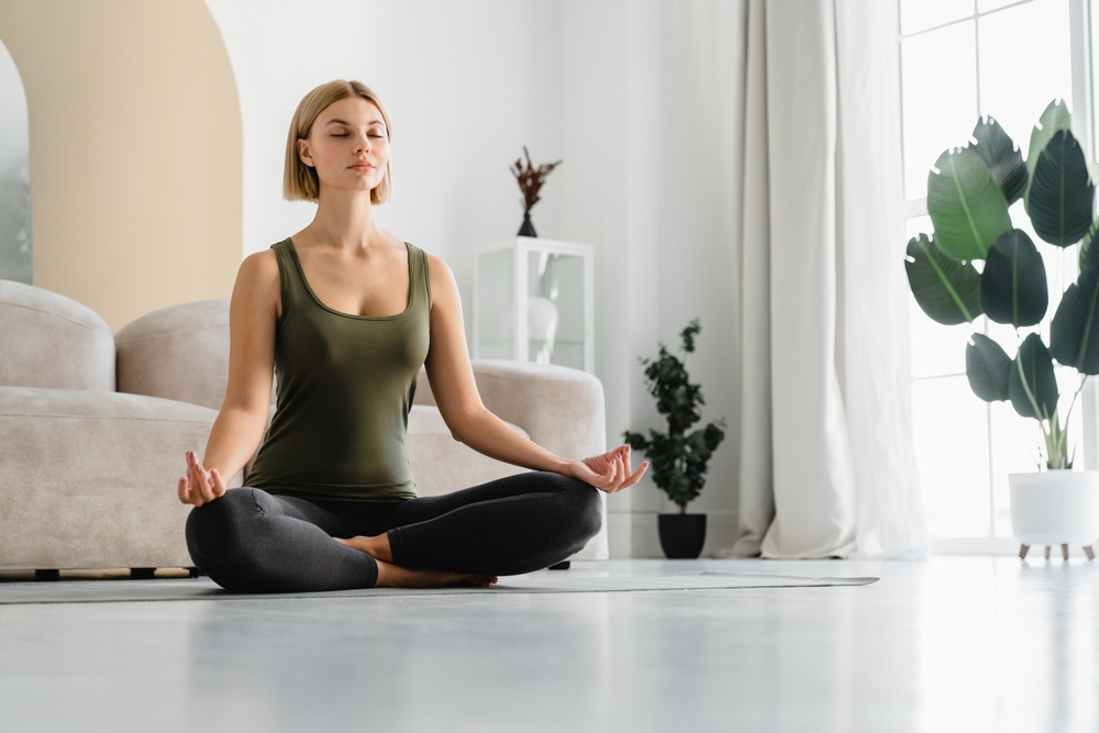 Relaxed young woman doing sitting in yoga position at home. Female athlete sitting in lotus meditating. Serene people concept. Life balance
