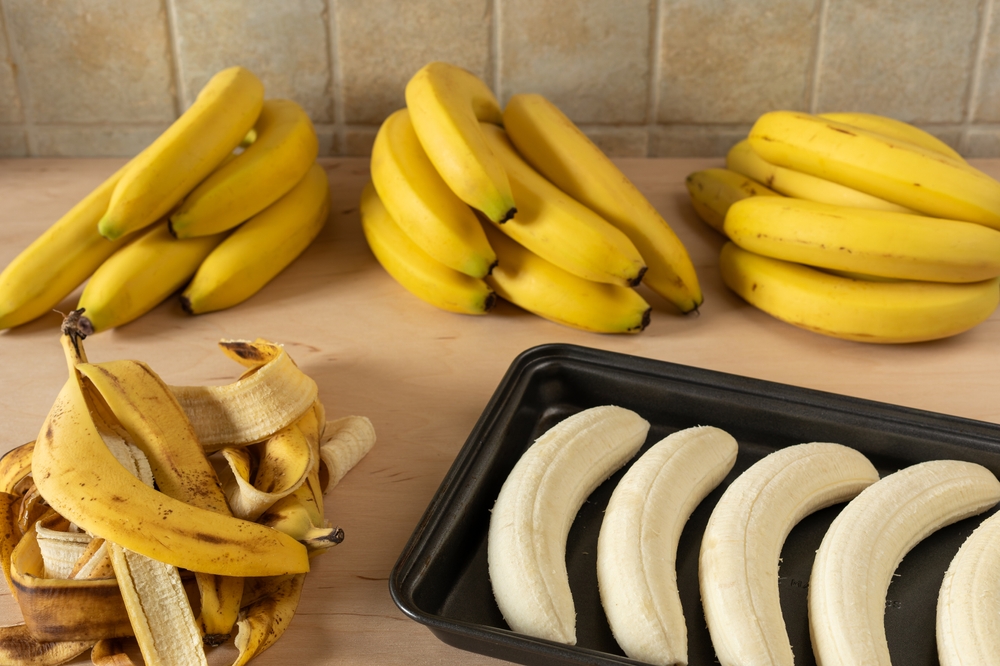 Preparing baked bananas - healthy, sweet fruit dessert. Baking tray with peeled bananas, a heap of banana peel, and three bunches of unpeeled bananas behind.
