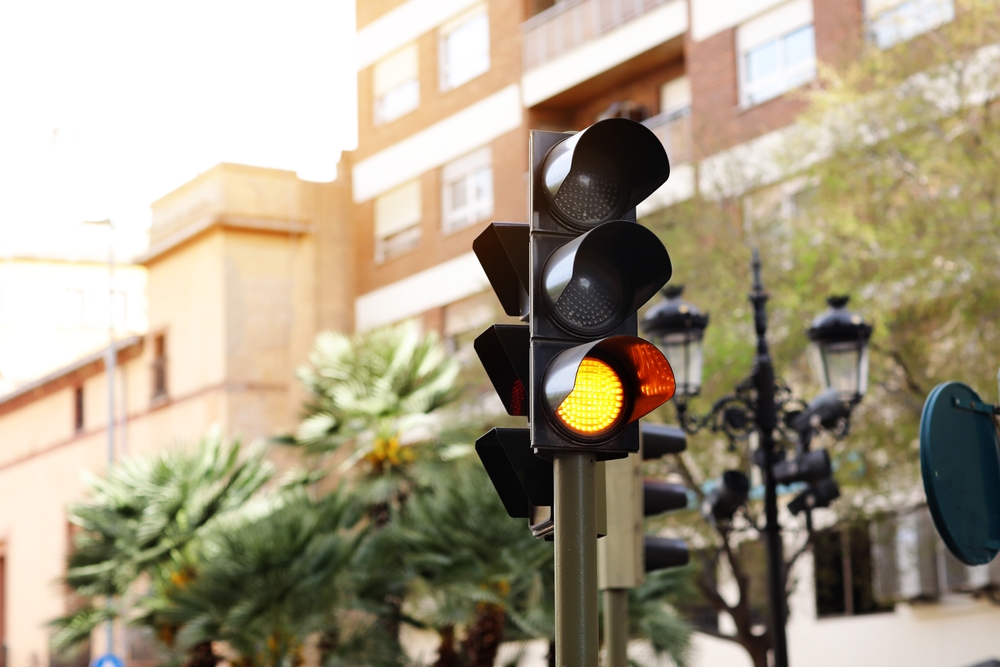 LED yellow traffic light on defocused european buildings and palms trees background. Attention. City crossing with amber light in semaphore, traffic control and regulation concept. Valencia, Spain.
