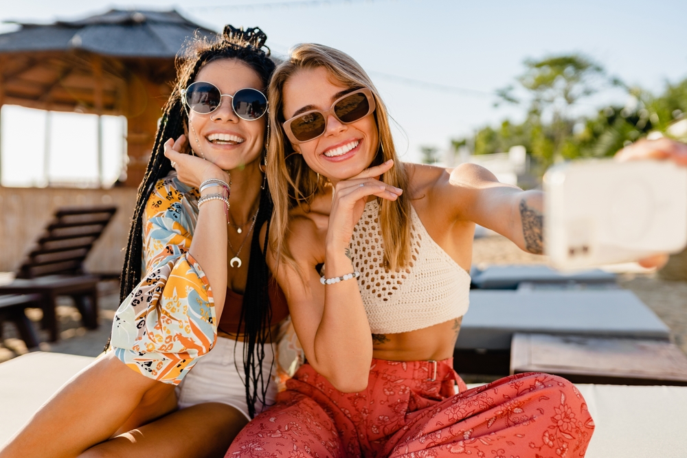 stylish smiling beautiful women relaxing having fun on beach in summer fashion trend outfit, colorful apparel boho style, tropical vacation wearing sunglasses, using smartphone taking selfie picture
