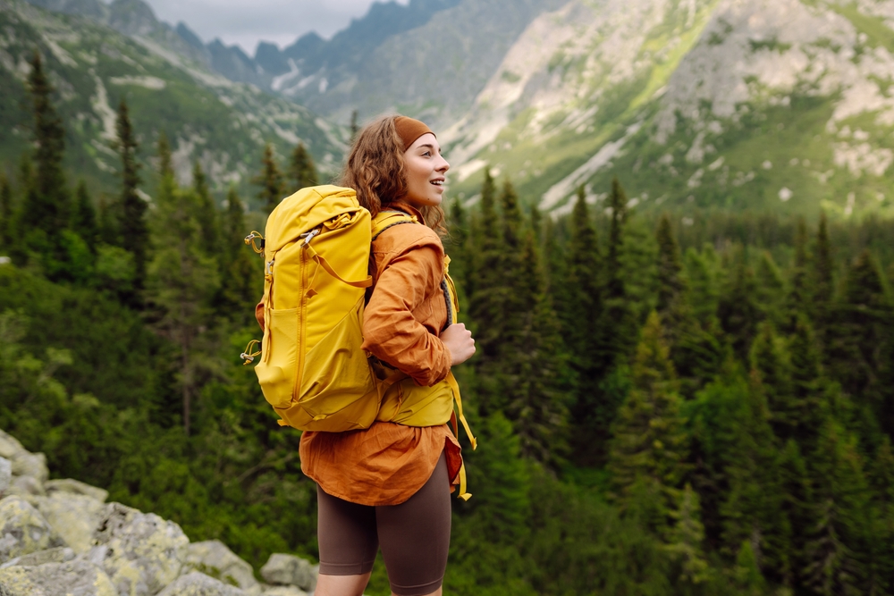 A smiling female traveler with a yellow hiking backpack enjoys the beautiful scenery of the majestic mountains. Travel, adventure. Concept of an active lifestyle.
