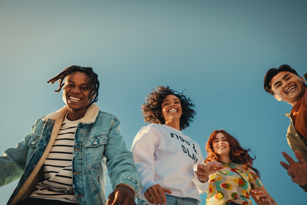 Group of happy young friends having fun outdoors, enjoying sunny day under blue sky. Joyful Gen Z people embracing friendship and laughter.