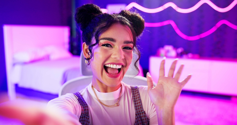 Selfie, influencer and girl with live streaming update in neon bedroom for social media audience, fans or followers. Face of excited content creator, gamer or streamer with wave hello in virtual post