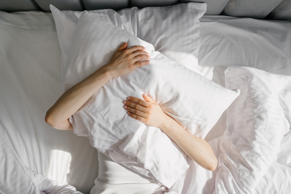 Female cover her head with cushion suffers from insomnia need sleeping pills, flat lay shot. Top above view of woman hug pillow, feeling deep sense of hopelessness lying on bed in bedroom
