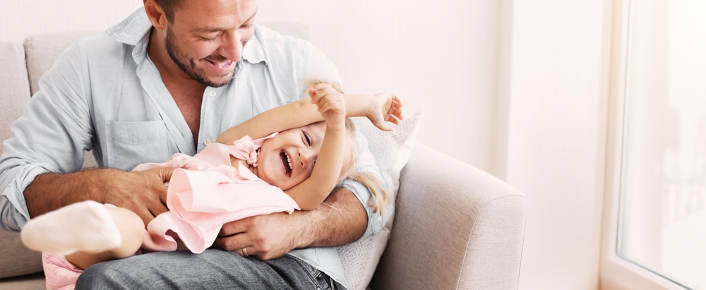 Best Dad In The World. Millennial man tickling his daughter who lying on his lap on the sofa. Free space