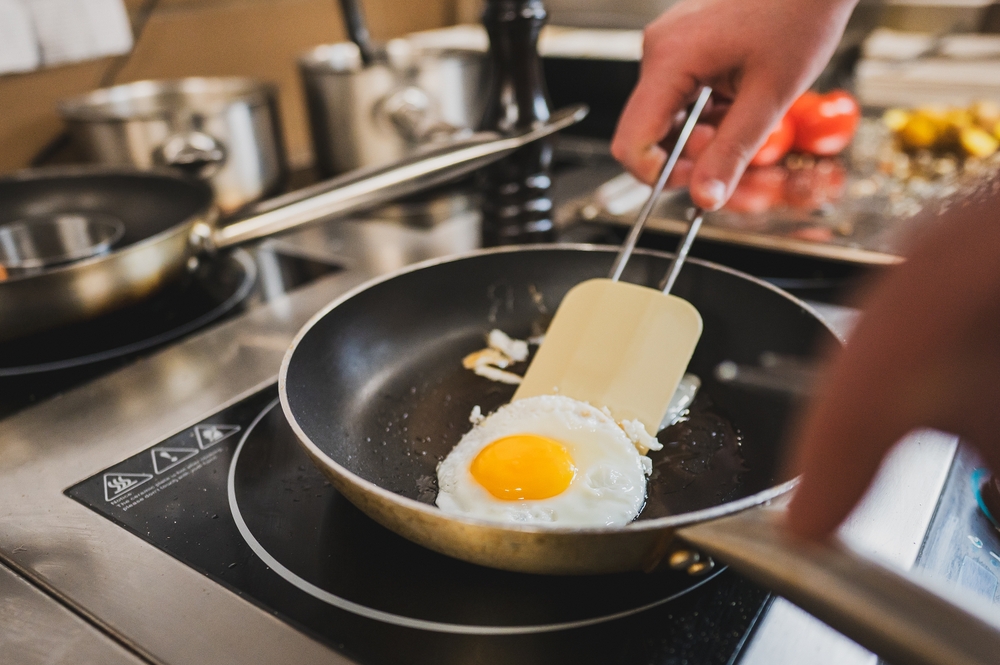 A close-up of a chef's hand using a spatula to flip a sunny-side-up egg in a frying pan. The kitchen setting features pots and pans in the background, showcasing a cooking scene.