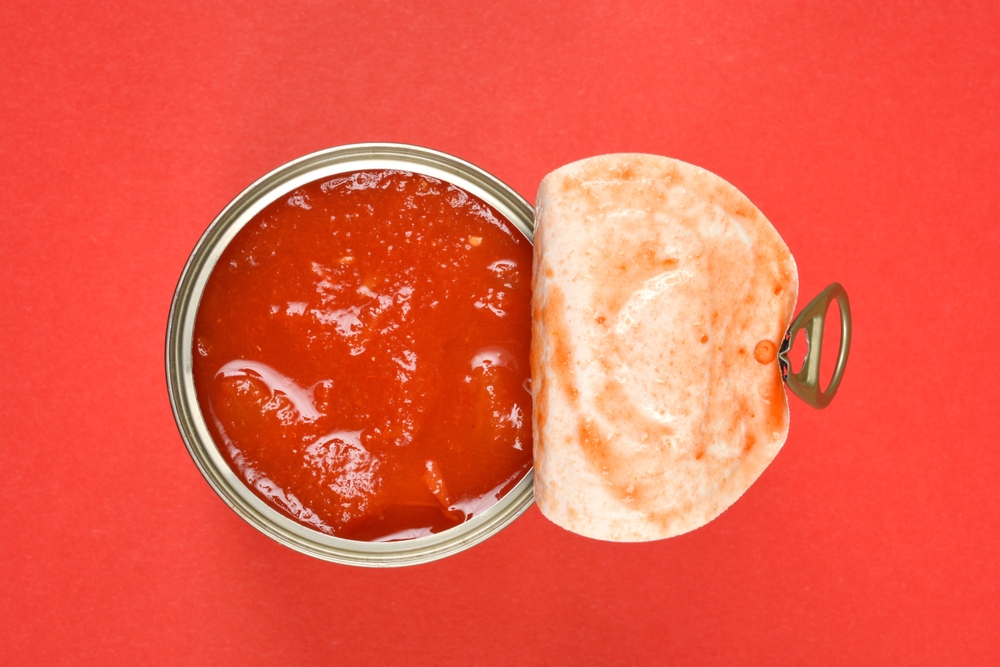 A top-down view shows an open can of chunky red tomato sauce with its lid peeled back, set against a vibrant red background
