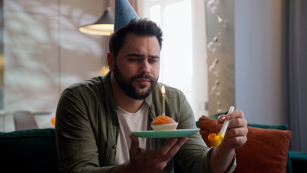 Depressed lonely sad despair Caucasian business man male guy businessman homeowner client festive cap head celebrating birthday alone holding plate with burning candle cupcake blowing whistle at home