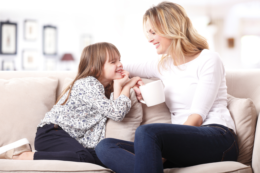 Portrait of a beautiful mother and her little girl sitting at home and sharing a happy moment together. Blond mom holding in her hand a cup of tea.