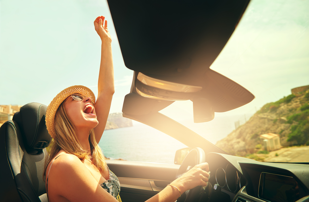 Excited pretty woman with hat and sunglasses reaching her fist in air as if to celebrate in convertible top automobile
