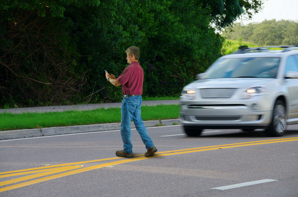 A man who is completely distracted by his cell phone is unaware that he is jaywalking by walking out into heavy traffic on a busy highway road.
