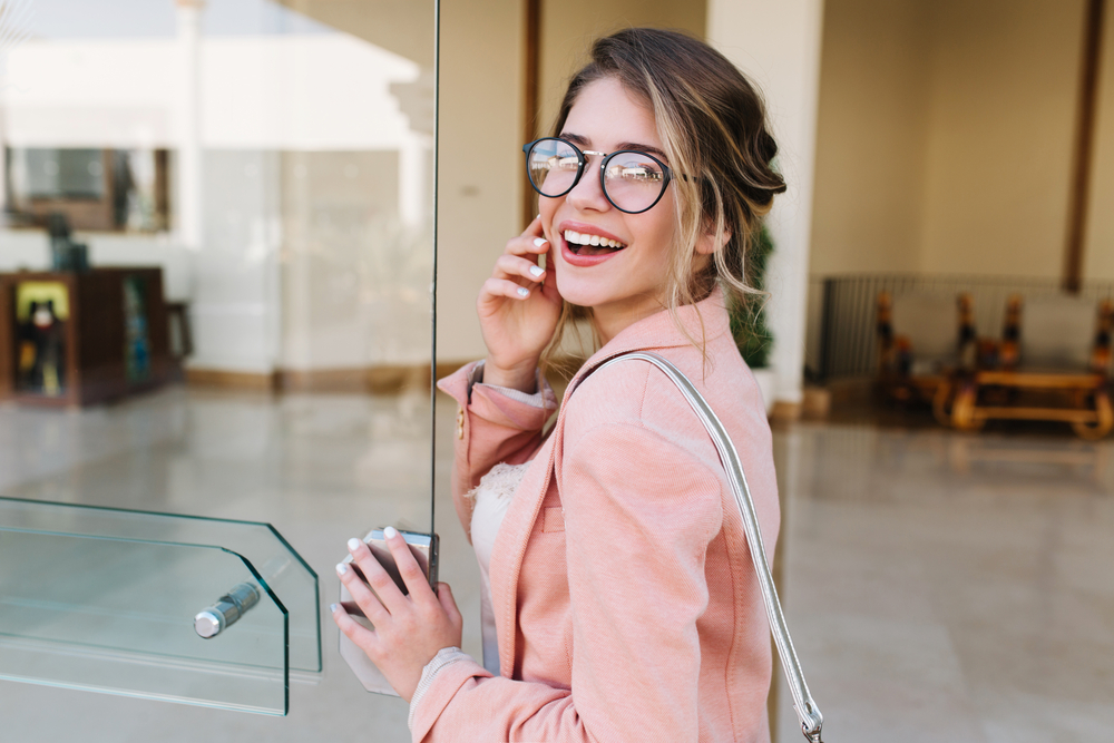 Closeup portrait of pretty young woman with hair up entering building, glass door into office, hotel, business centre. Wearing fashionable glasses, gray pants, pink jacket.