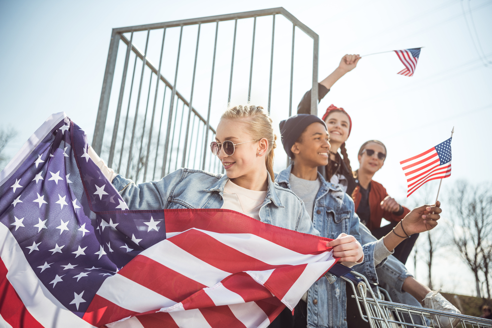 Happy teenagers group having fun and waving american flags at sunset