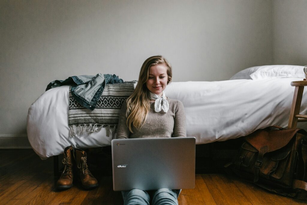woman sitting beside a bed while using a laptop
