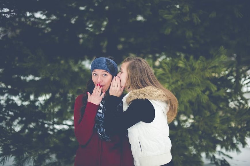 A woman swhispering into another womans ear, with a looks of surprise on her face.