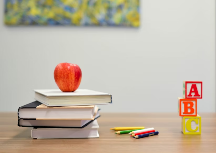 school books stack with an apply on top, coloured pencils and ABC blocks. 