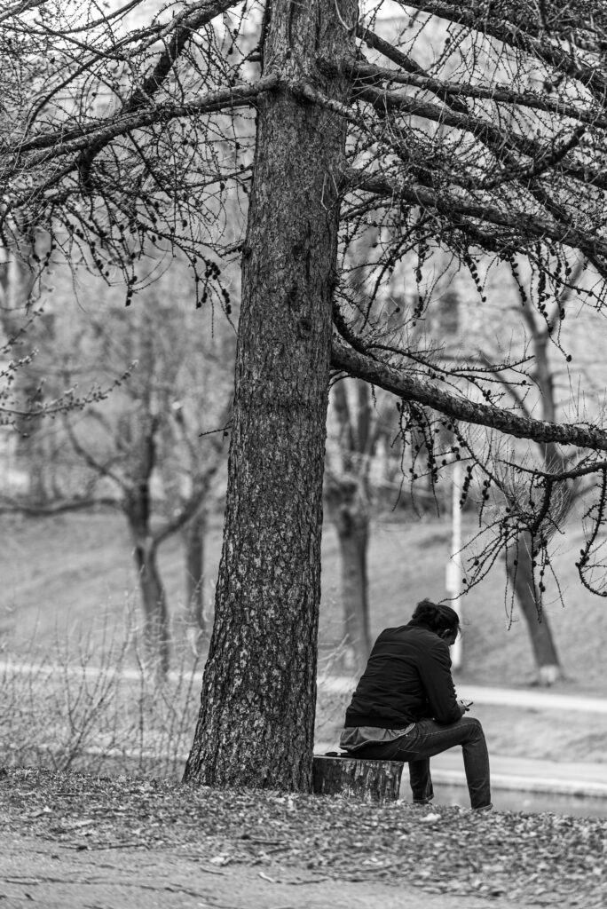 man in black jacket sitting on bench near bare trees during daytime
