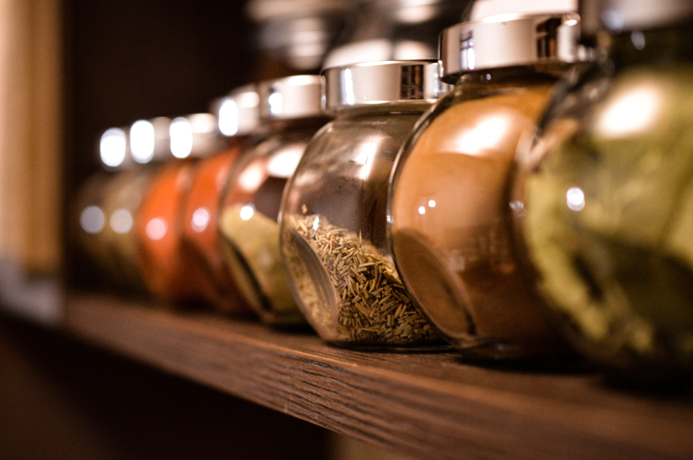 Spice jars on a shelf in kitchen being displayed inline, paprika, pepper, spices, rosemary, basil, curry. focus is done on rosemary.
