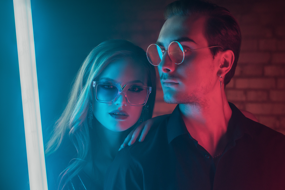 Fashion portrait of couple: beautiful woman and handsome man posing in colorful bright neon uv blue and red lights. Models wearing trendy glasses. Club, disco style