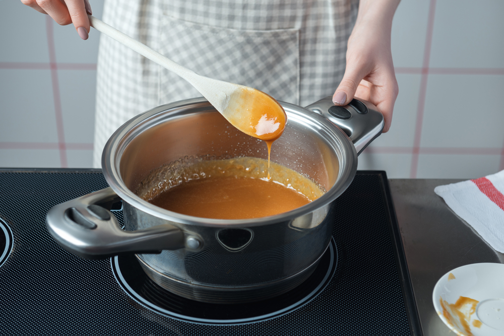 Women in checkered apron with wooden spoon preparing caramel in the stainless steel pan.

