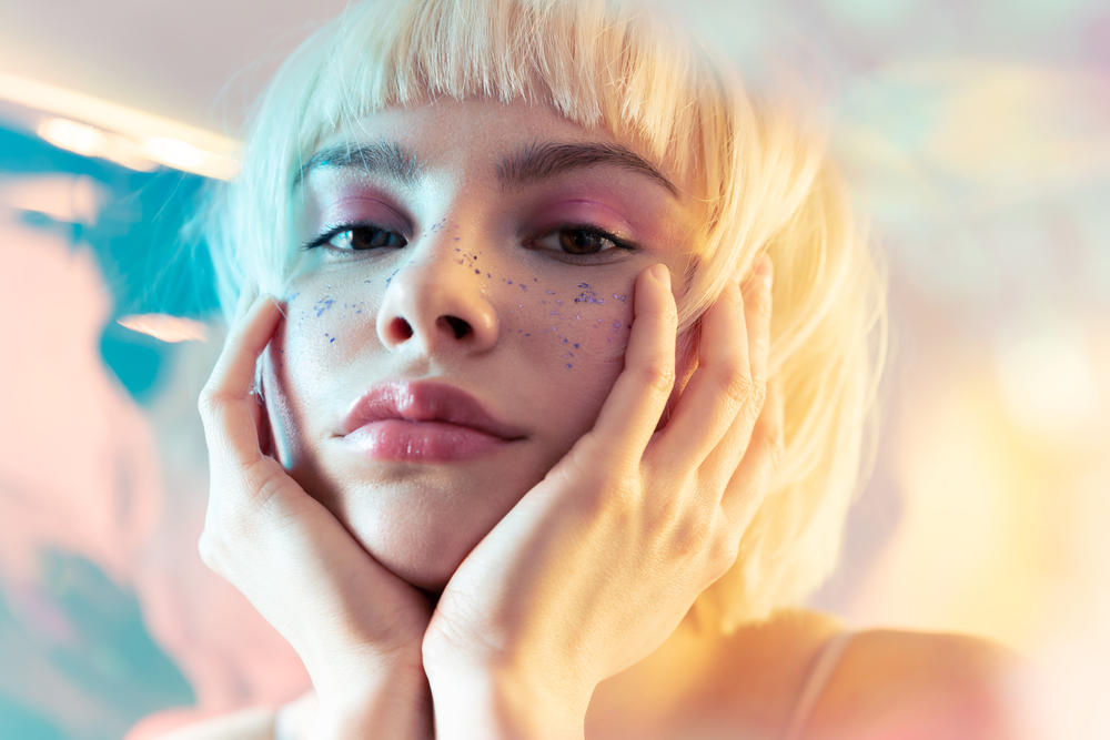 Young pensive blond teen girl with perfect brows, smooth skin and modern makeup touch cheeks with hands. Pretty fashion model with short haircut and sparkles on face. Beauty salon advertising photo