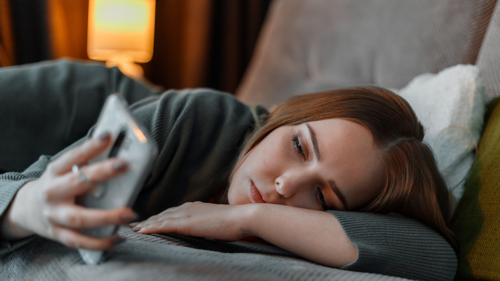 Sad teenage girl suffering from insomnia surfing Internet or chatting using smartphone at night lying on bed. Depression Internet addiction and depression in young woman. Long web banner.
