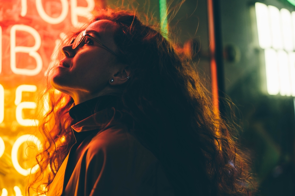 Curly haired young woman tourist with light makeup in glasses looks around standing near bar with colorful neon sign against night megalopolis