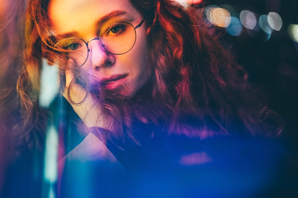 Curly haired young woman standing near bar with colorful neon sign against night megalopolis