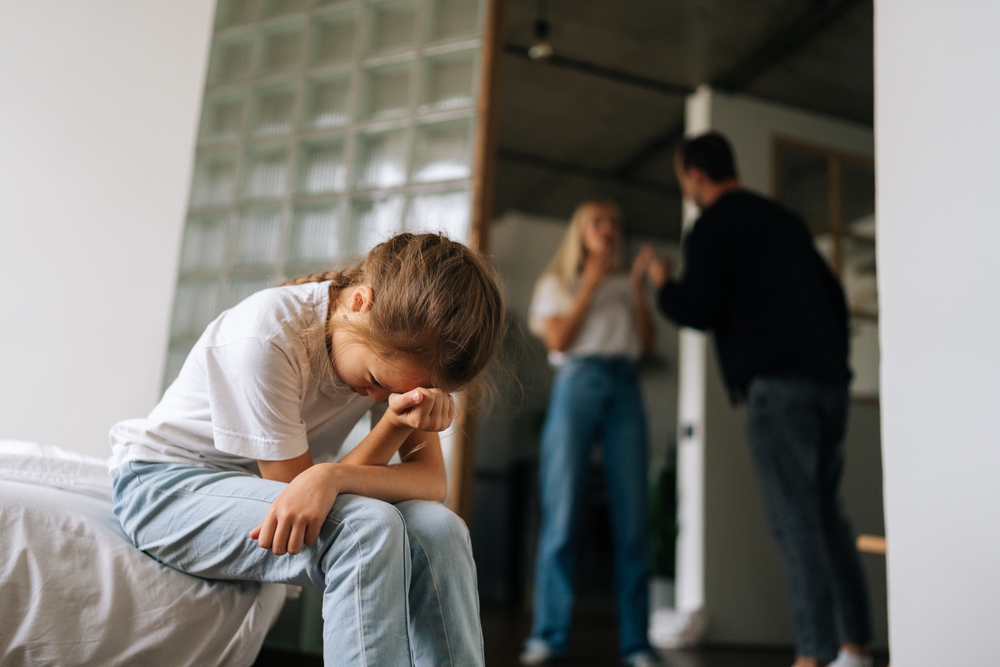 Low-angle view of despair lonely little girl crying suffering sitting on sofa during parents quarrelling and fighting in living room on background. Concept of family problems, conflict, crisis.