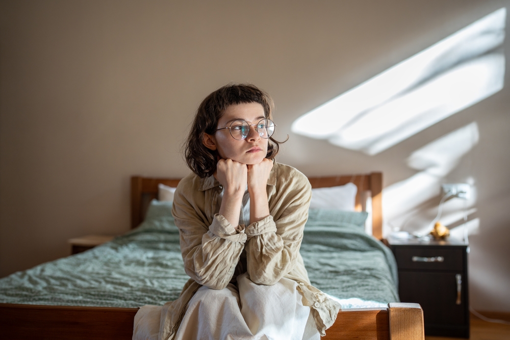 Depressed lonely woman sitting on cozy bed sadly look at window feeling anxiety. Alone girl suffering from emotional pain having life troubles, problems. Mental disorder anhedonia, stress discomfort