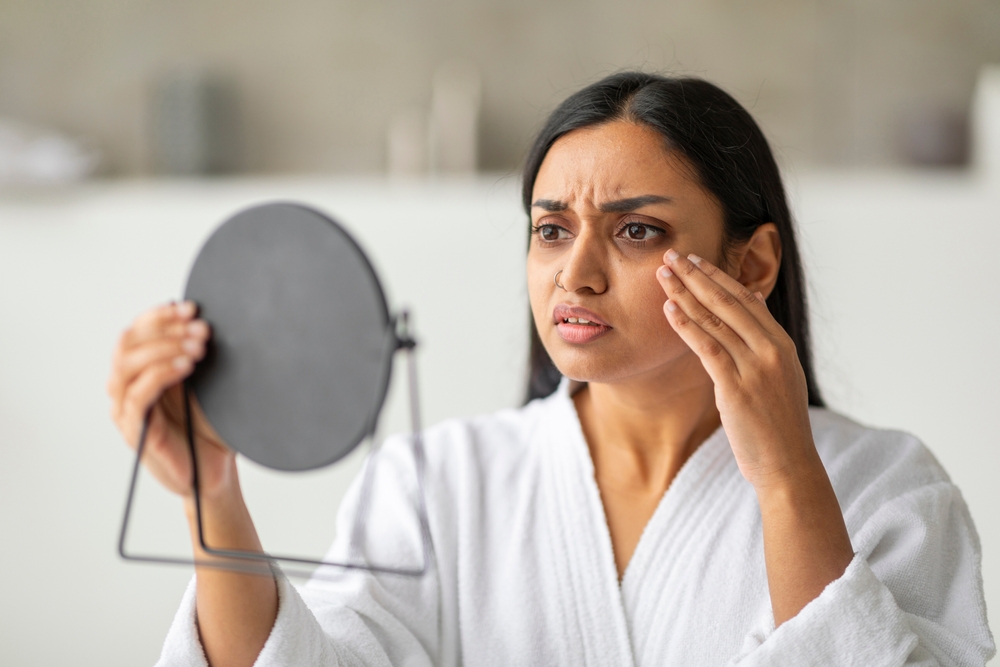 Upset young indian woman in white bathrobe looking at mirror and touching her cheeks, having oily or dry skin problem, puffy eyes in the morning, bathroom interior, closeup photo
