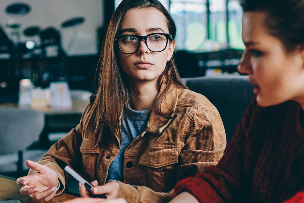 Selective focus on serious hipster girl in spectacles sitting college campus listening carefully female friend, two concentrated women studying together thinking on startup project using technology