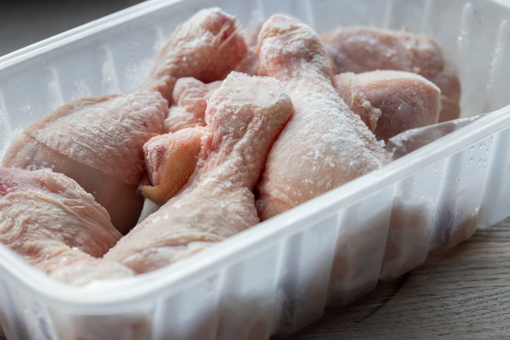 Frozen chicken drumsticks in a plastic container defrosting. On a wooden chopping board