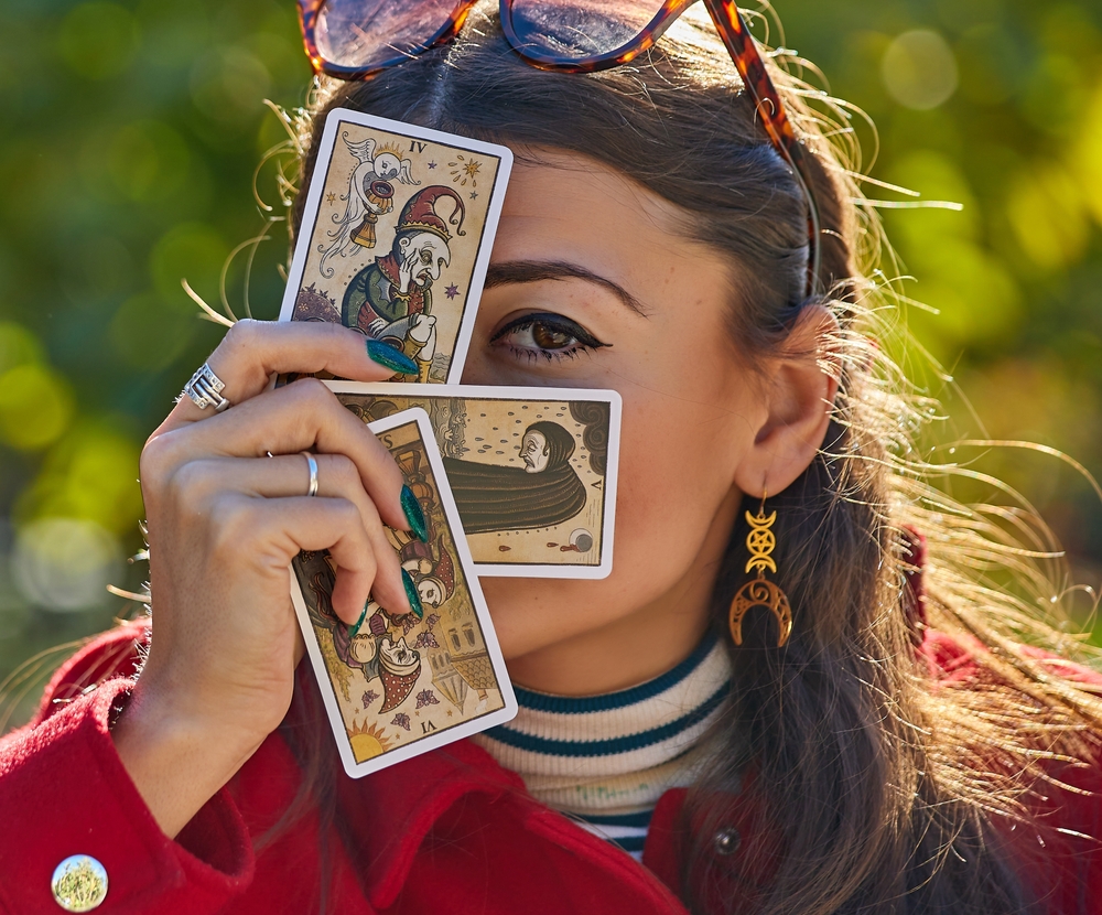 Almeria Spain - December 22,2024;A woman in a red coat holds three tarot cards in her hand, partially covering her face; she has green nails, multiple rings, and wears crescent moon earrings;